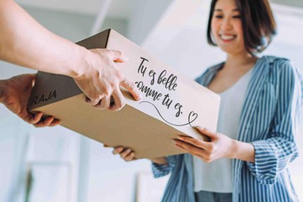 Eine lächelnde Frau erhält ein personalisiertes Paket mit der französischen Aufschrift: Tu es belle comme tu es.